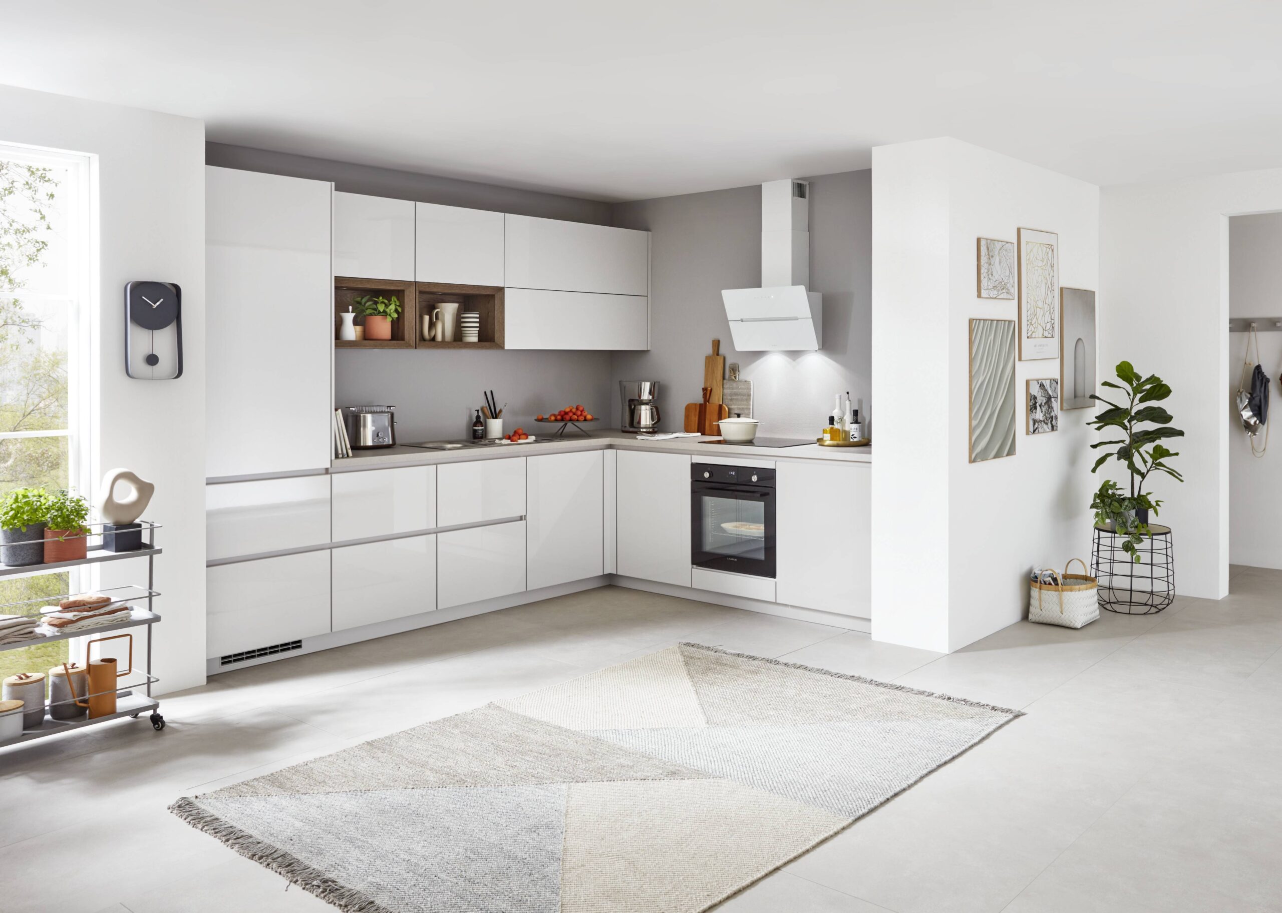 A white, modern L-shaped kitchen with integrated appliances, open shelving, and a geometric rug. A clock hangs on the wall.