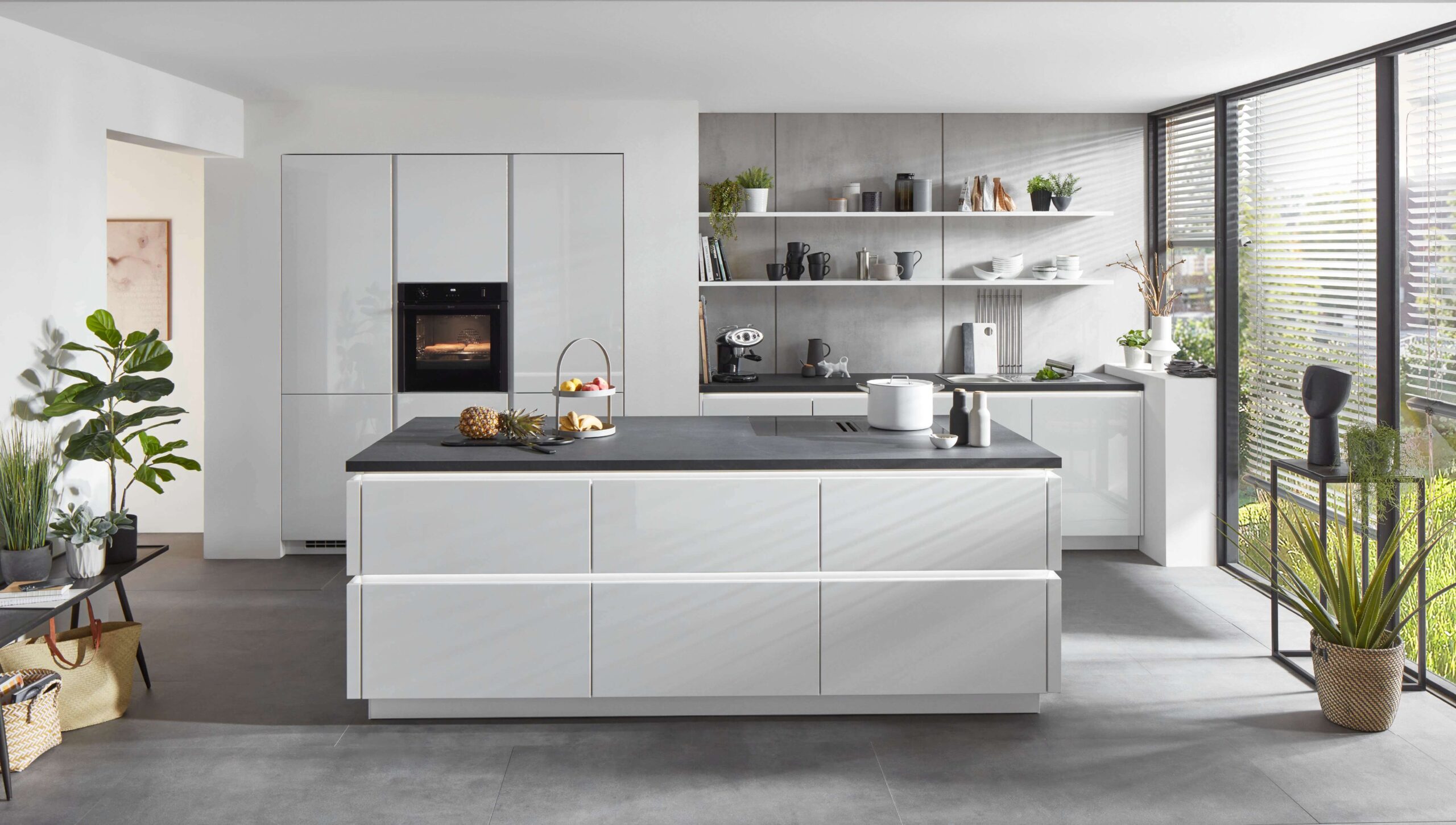 Sleek, modern white kitchen with a long black countertop island and open shelving. Large windows with blinds offer a view of greenery.