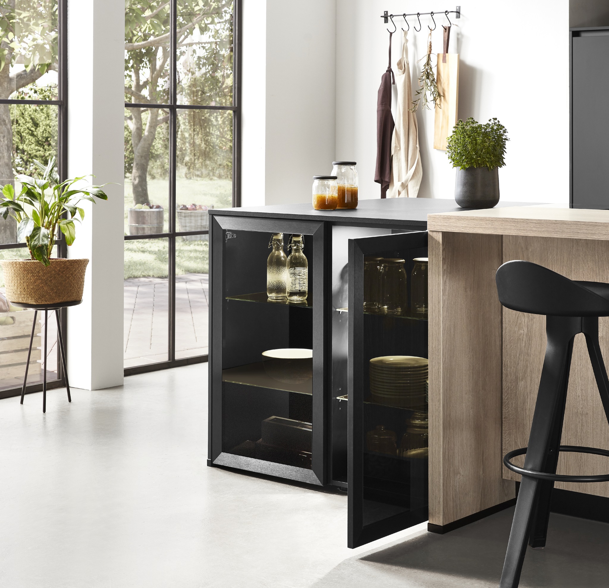 Close-up of a modern black kitchen island with open glass doors revealing dishware and storage. A black bar stool and large window are also visible.