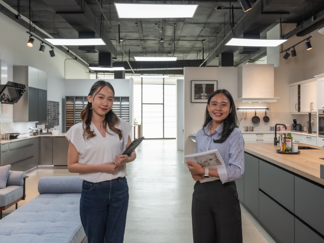 Two German Kitchen Design staff stand in a German Kitchen Design's showroom in River City Bangkok. One holds a tablet and the other a brochure.