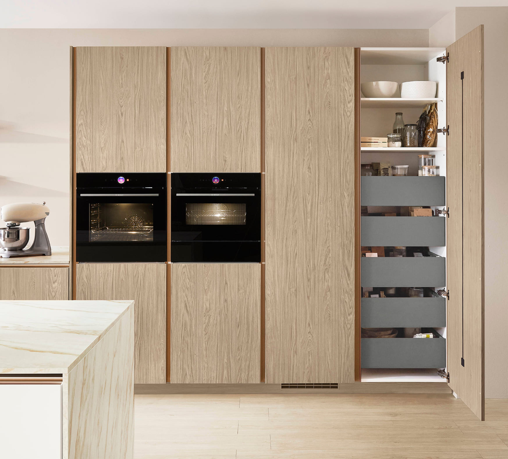 Modern kitchen with light wood cabinets, two black ovens, and an open pantry cabinet revealing gray drawers. A marble countertop is visible on the island.