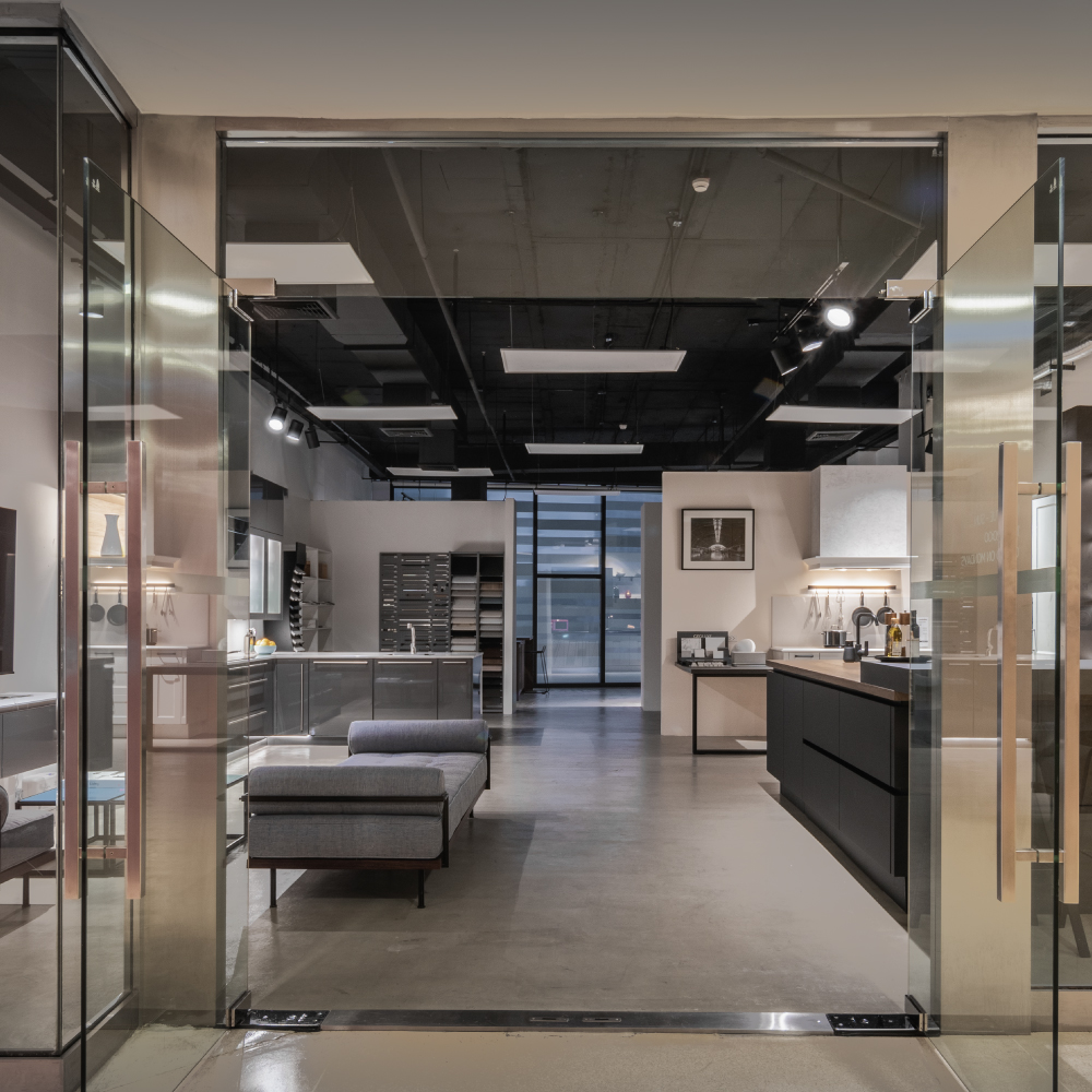 View through glass doors into German Kitchen Design's modern kitchen showroom with dark cabinets and a gray couch.