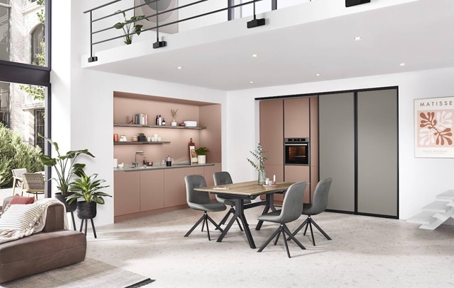 A stylish, open-plan high-end German kitchen featuring a unique pink and gray color scheme, a dining area, and large windows.