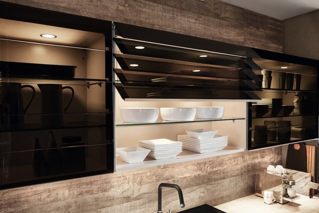 โฟกัสที่ตู้ครัวสไตล์โมเดิร์นพร้อมบานกระจก มีชั้นที่ส่องสว่างแสดงภาชนะสีขาว ดีไซน์ที่มีสไตล์และใช้งานได้จริงพร้อมพื้นผิวไม้ด้านหลัง