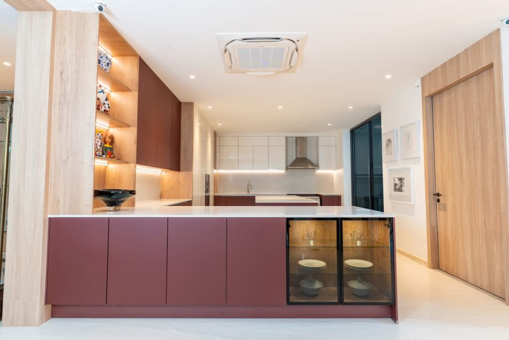 A minimalist kitchen design in a Bangkok condo featuring a rust red handleless island that acts as a furniture piece, seamlessly blending with the open-plan living area.