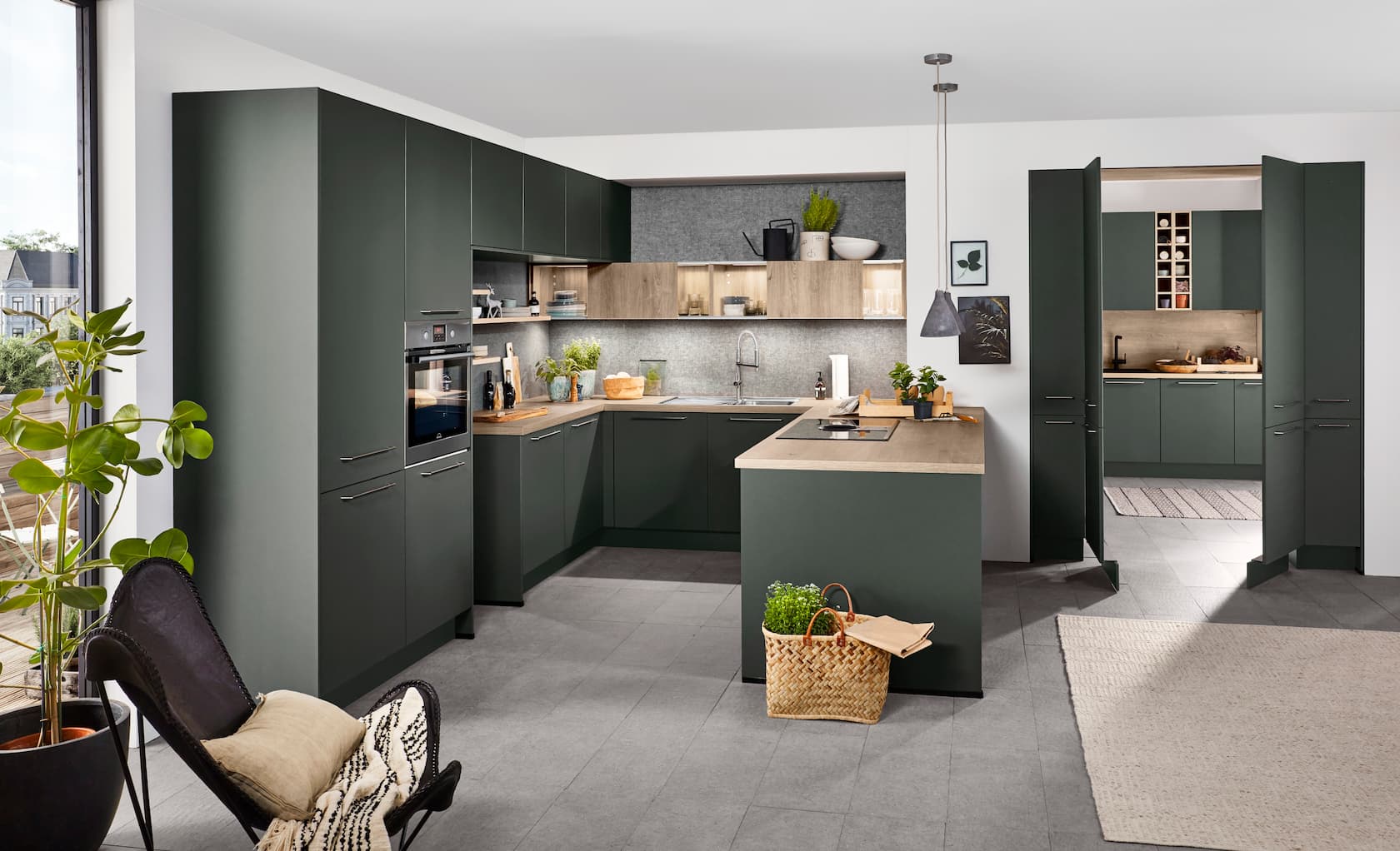 A modern U-shaped German kitchen showcasing matte mineral green cabinetry with subtle horizontal steel handles, paired with natural wood-grain countertops, matching open wooden shelving, and a built-in stainless steel oven.