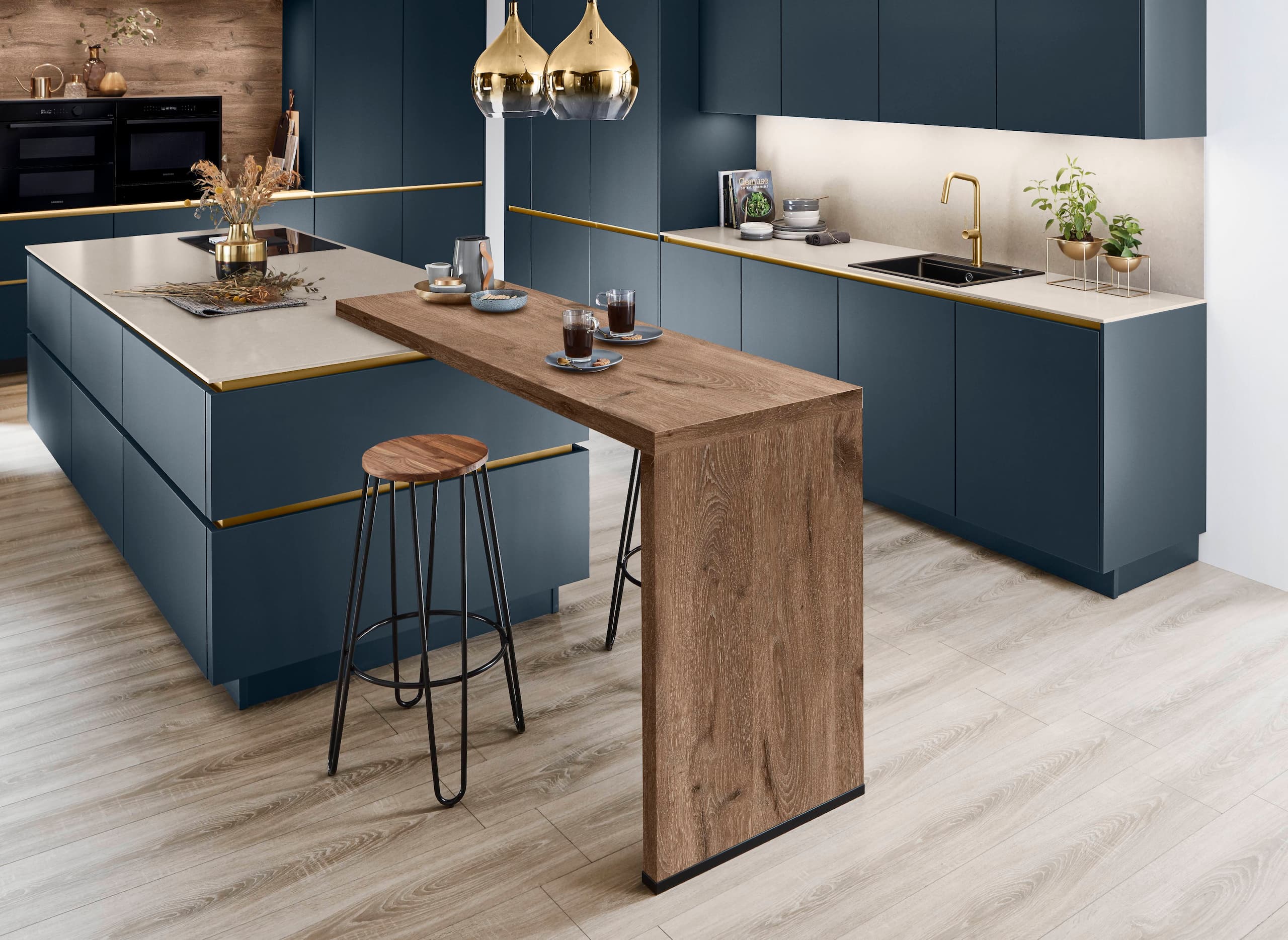 A luxurious handleless German kitchen featuring anti-fingerprint Fjord blue ultra matt cabinetry with elegant gold recessed handle tracks, a matching large central island with an attached elevated raw wood breakfast bar, premium gold plumbing fixtures, and dual integrated ovens.