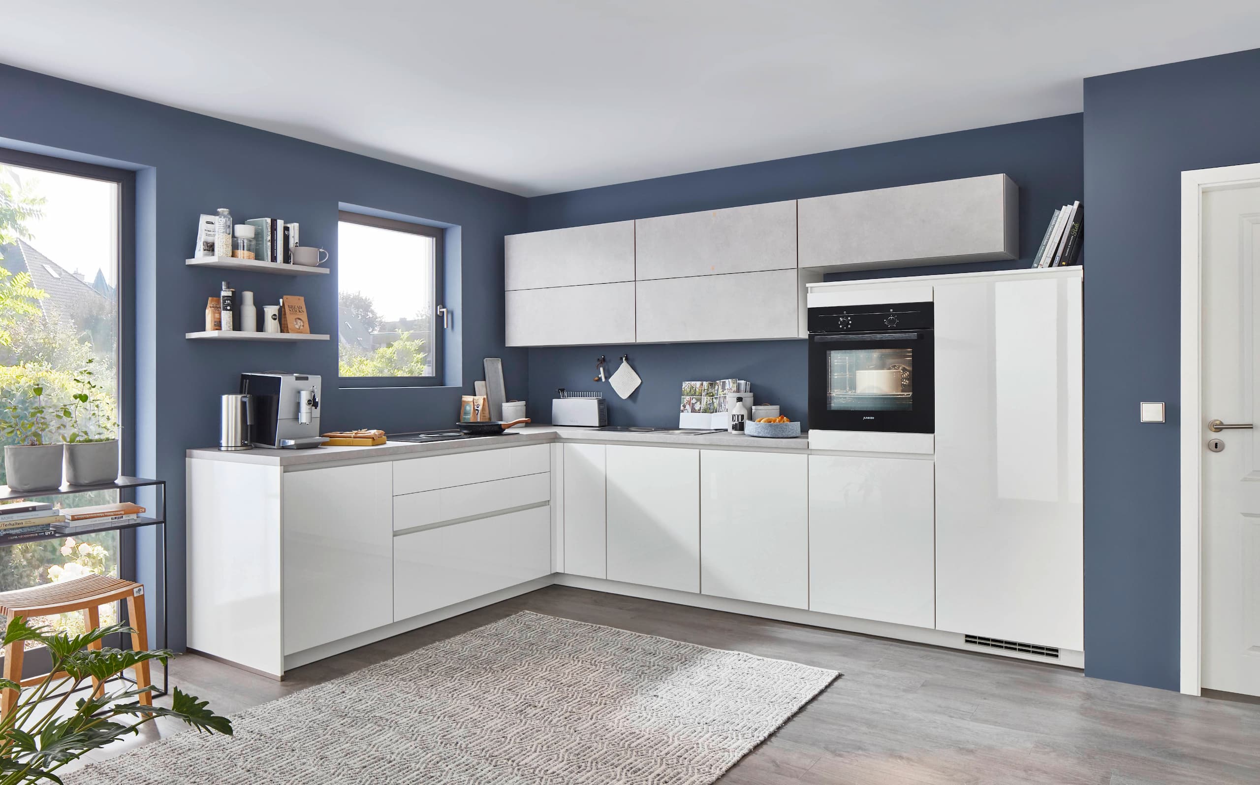 A contemporary handleless L-shaped German kitchen showcasing alpine white high gloss base units and tall appliance housing, paired with textured concrete-look grey upper wall cabinets, set against a deep blue feature wall with an integrated black oven.