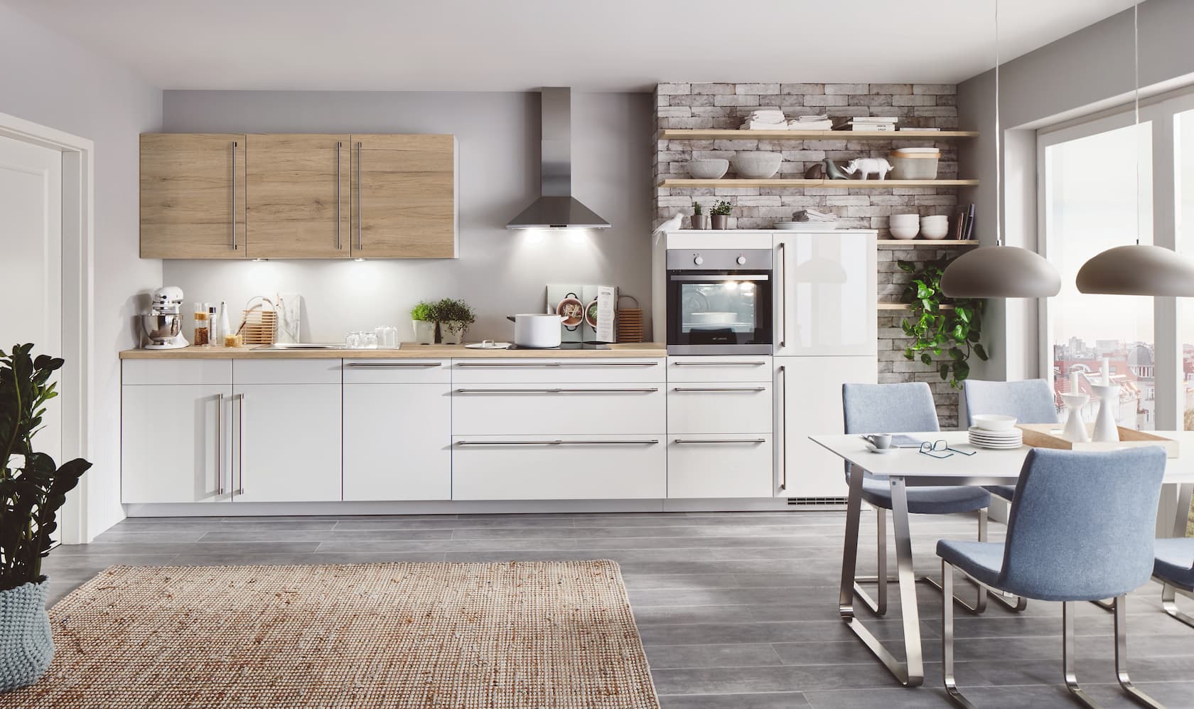 A bright modern German kitchen featuring high-gloss white base cabinets with long stainless steel bar handles, light wood-grain upper cabinetry, a matching wood-texture countertop, and an exposed brick accent wall with open wooden shelving.