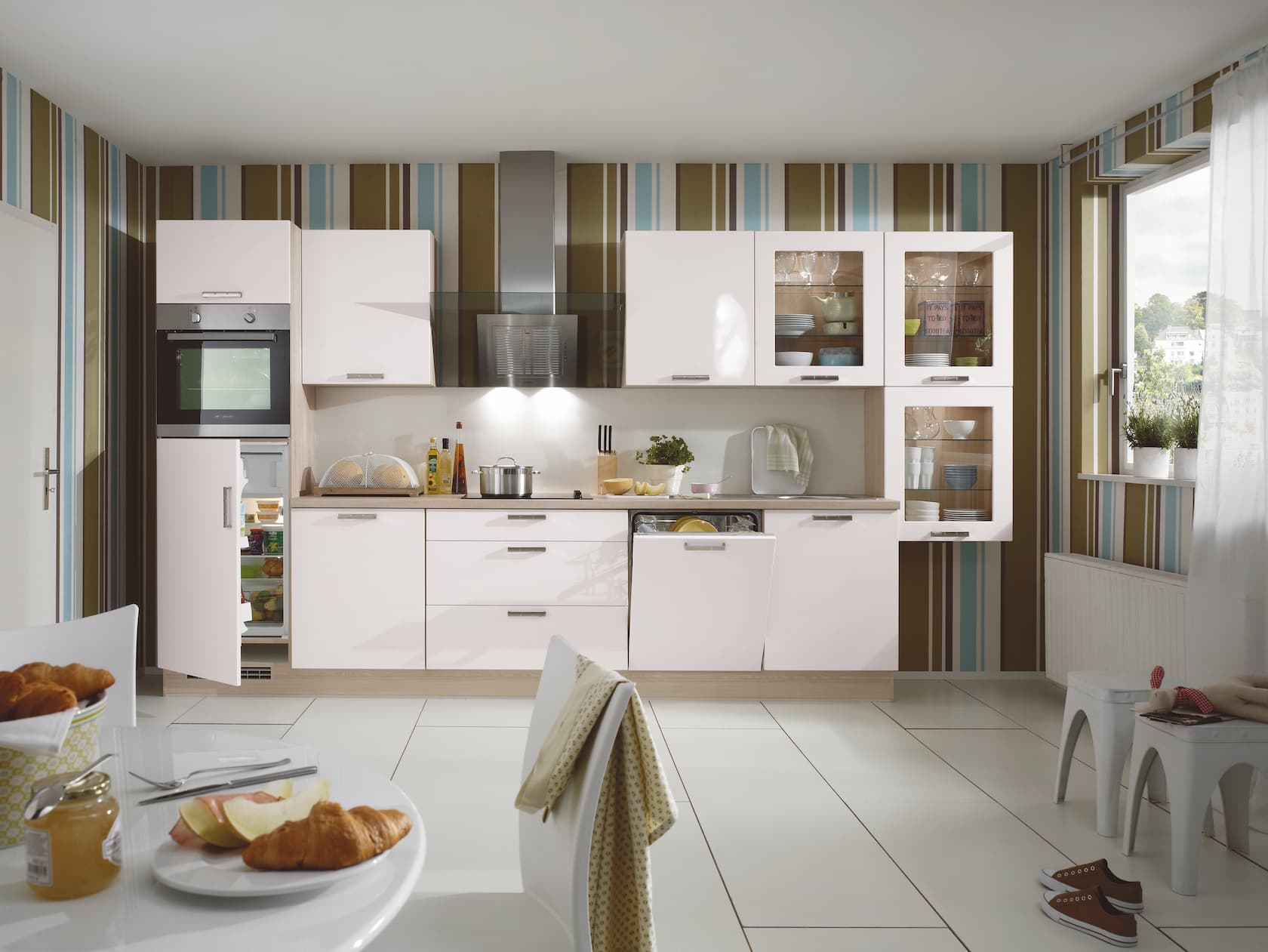 A modern ultra high-gloss alpine white German kitchen featuring sleek stainless steel bow handles, glass-front upper display cabinets, integrated appliances including an open dishwasher and elevated oven tower, and a light wood-grain countertop and framing.