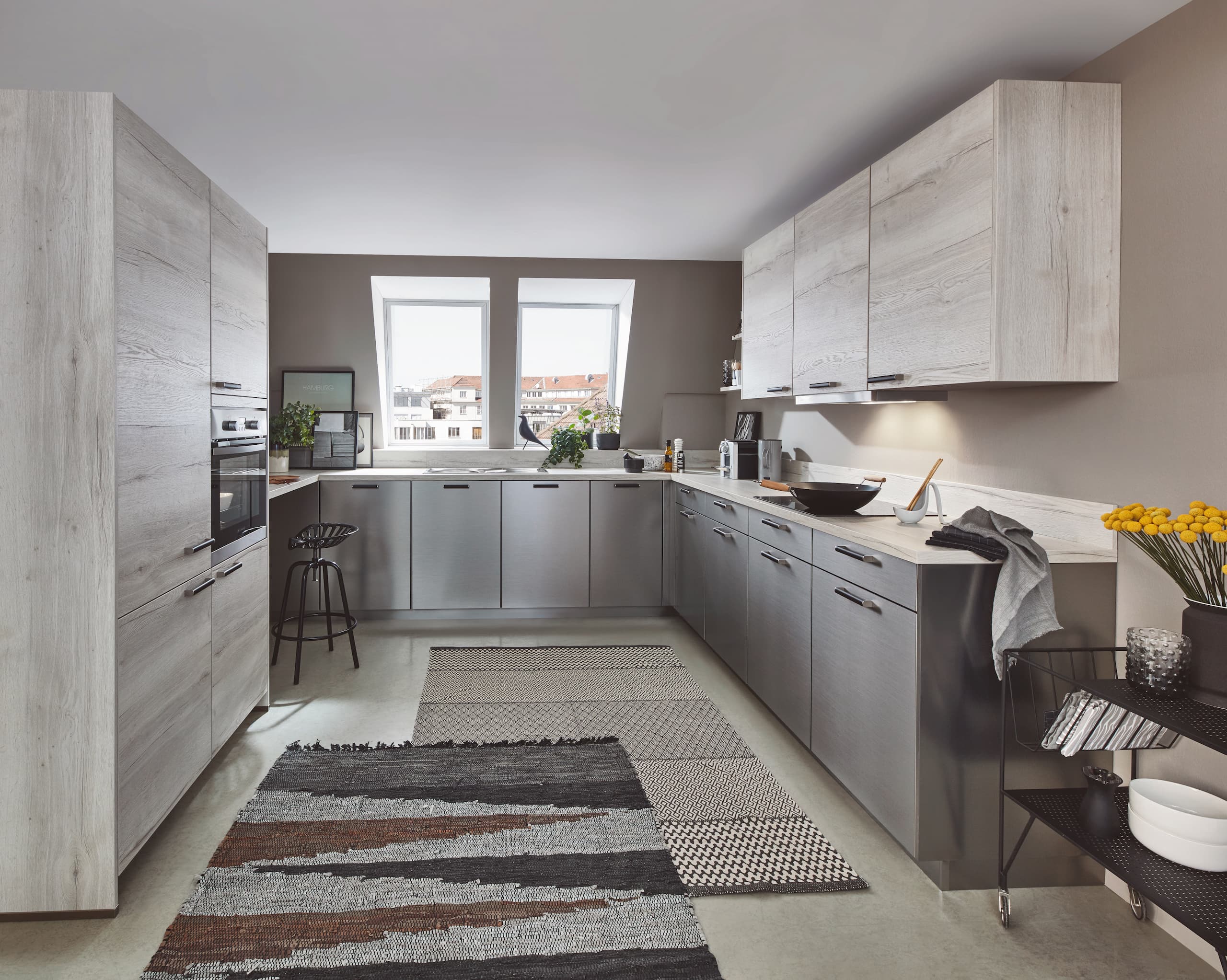 A modern U-shaped German kitchen showcasing a brushed stainless steel reproduction finish on the base units, paired with light, concrete-grey wood grain tall storage towers and upper wall cabinets, complete with slim dark horizontal handles.
