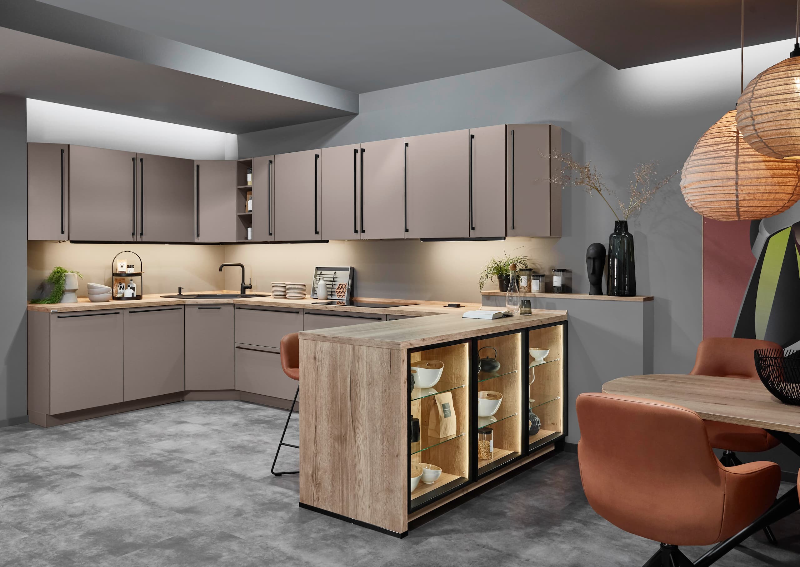 A contemporary U-shaped German kitchen showcasing smooth Macchiato-colored cabinetry with sleek vertical black bar handles, paired with a warm Sierra oak reproduction kitchen island that features an illuminated open glass-shelf display and integrated accent lighting.