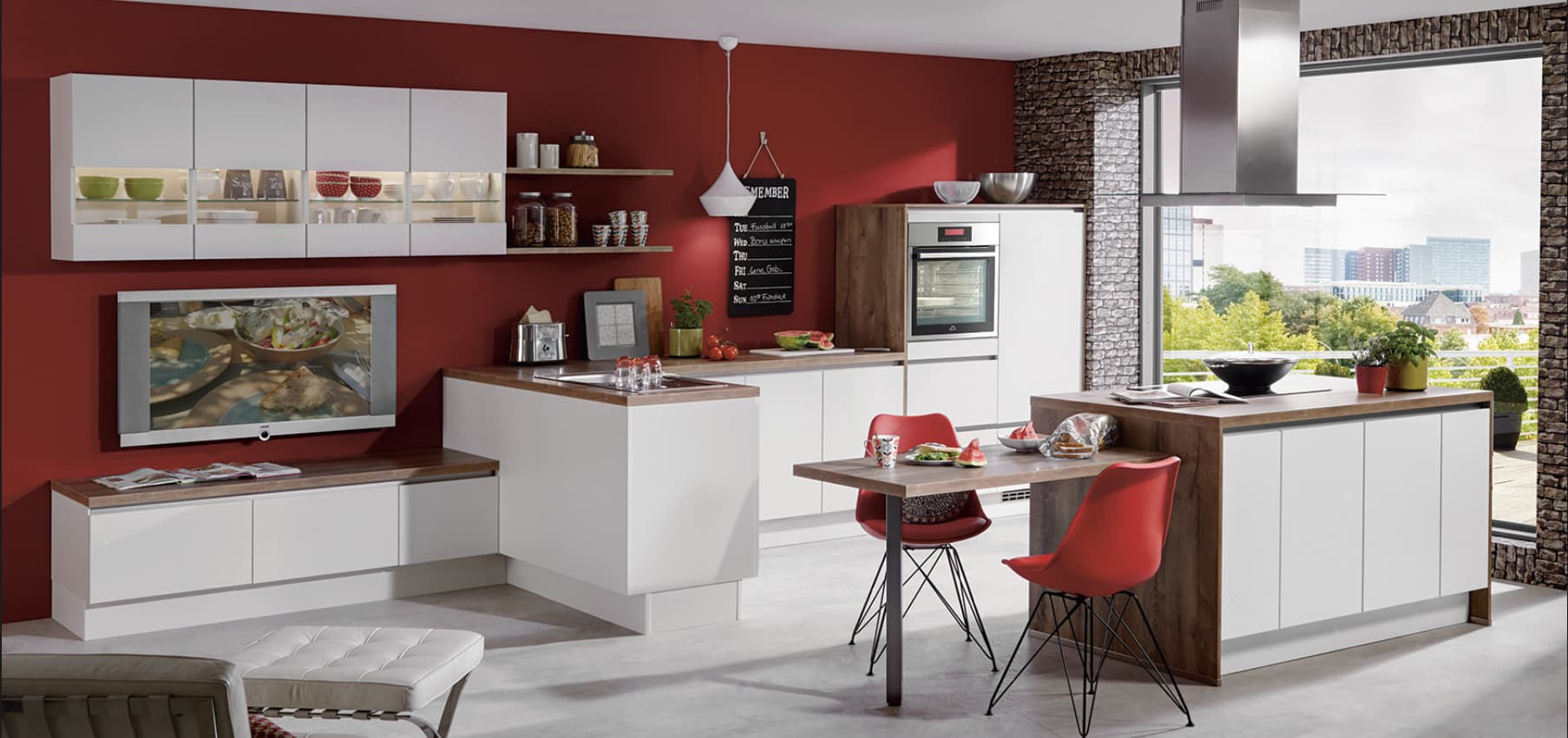 A contemporary handleless open-plan German kitchen featuring clean white matte cabinetry, warm natural wood-grain countertops and side cladding, an integrated kitchen island with an attached matching wood dining table, and a row of illuminated glass-bottom upper wall cabinets against a deep red feature wall.