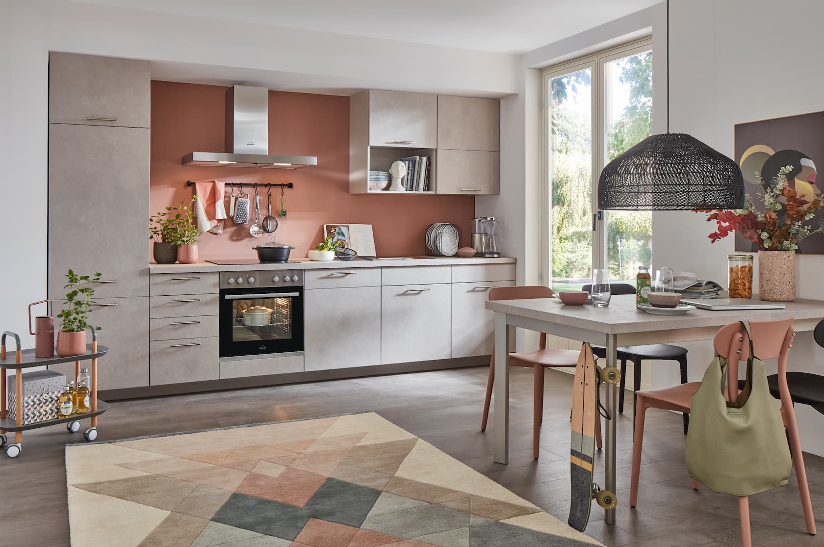 A modern single-wall German kitchen featuring textured concrete sand reproduction cabinetry with slim stainless steel handles, set against a warm terracotta accent wall, paired with an integrated stainless steel extractor hood and a matching concrete-look dining table setup.