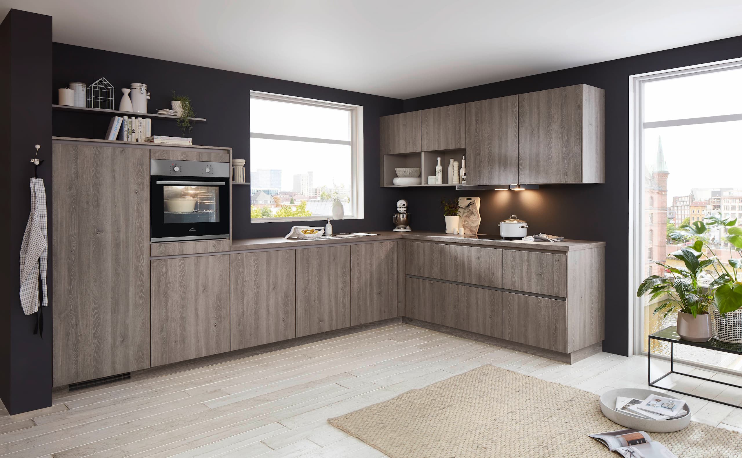 A modern handleless L-shaped German kitchen showcasing textured Bergamo oak reproduction cabinetry with seamless recessed handle profiles, a matching slim wood-grain worktop, upper wall units with an integrated under-cabinet light, and an elevated integrated silver oven set against a matte black wall.