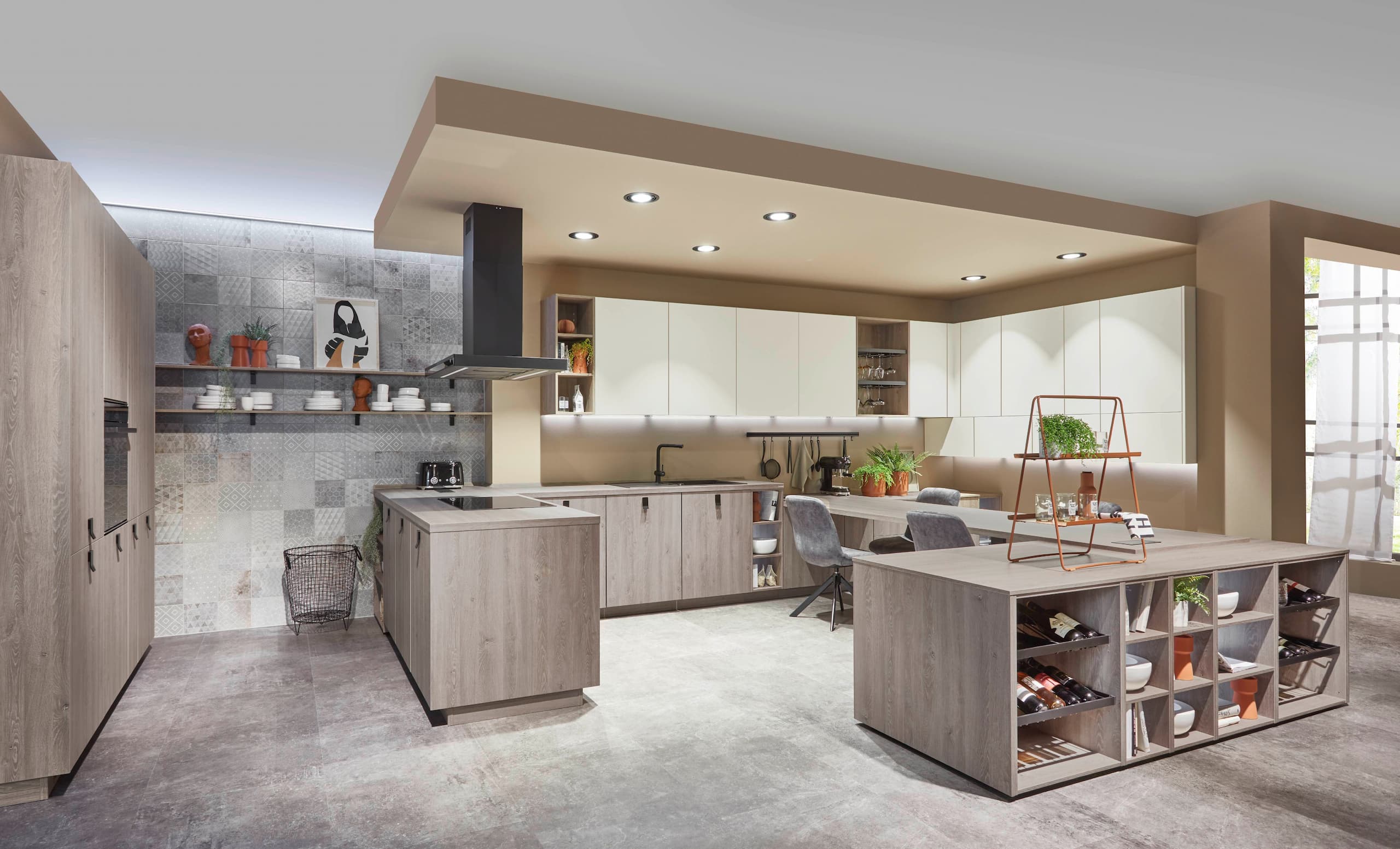 A spacious, modern German kitchen showcasing textured Bergamo oak reproduction base units and storage towers with minimalist vertical black handles, contrasted by matte sand-colored upper cabinetry, a matching drop-ceiling panel with integrated spotlights, and a large multi-compartment open wine rack island.