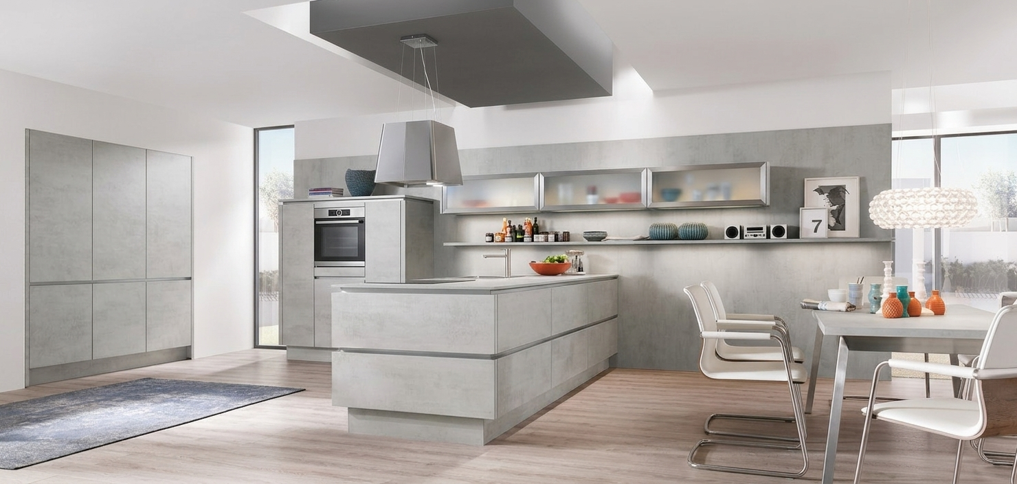 A minimalist handleless German kitchen showcasing a seamless concrete grey reproduction finish on the tall cabinetry, matching central kitchen island, and rear wall cladding, accented by horizontal glass-front lift-up wall cabinets, an integrated stainless steel wall oven, and a modern floating box extractor hood.