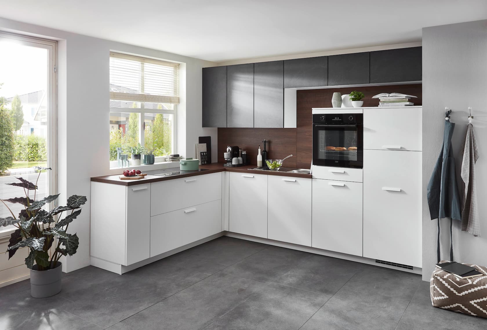 A modern L-shaped German kitchen featuring matte white base cabinets and tall appliance housing with matching white handles, contrasted by dark slate-grey upper cabinets, a rich dark wood-grain countertop and matching backsplash, and an integrated black oven.