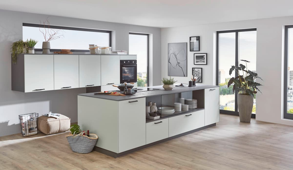 A modern German kitchen featuring matte honed jade green cabinetry with sleek black handles, a dark grey stone-texture kitchen island with integrated open shelving, and a built-in black oven in a bright, minimalist open-plan space.