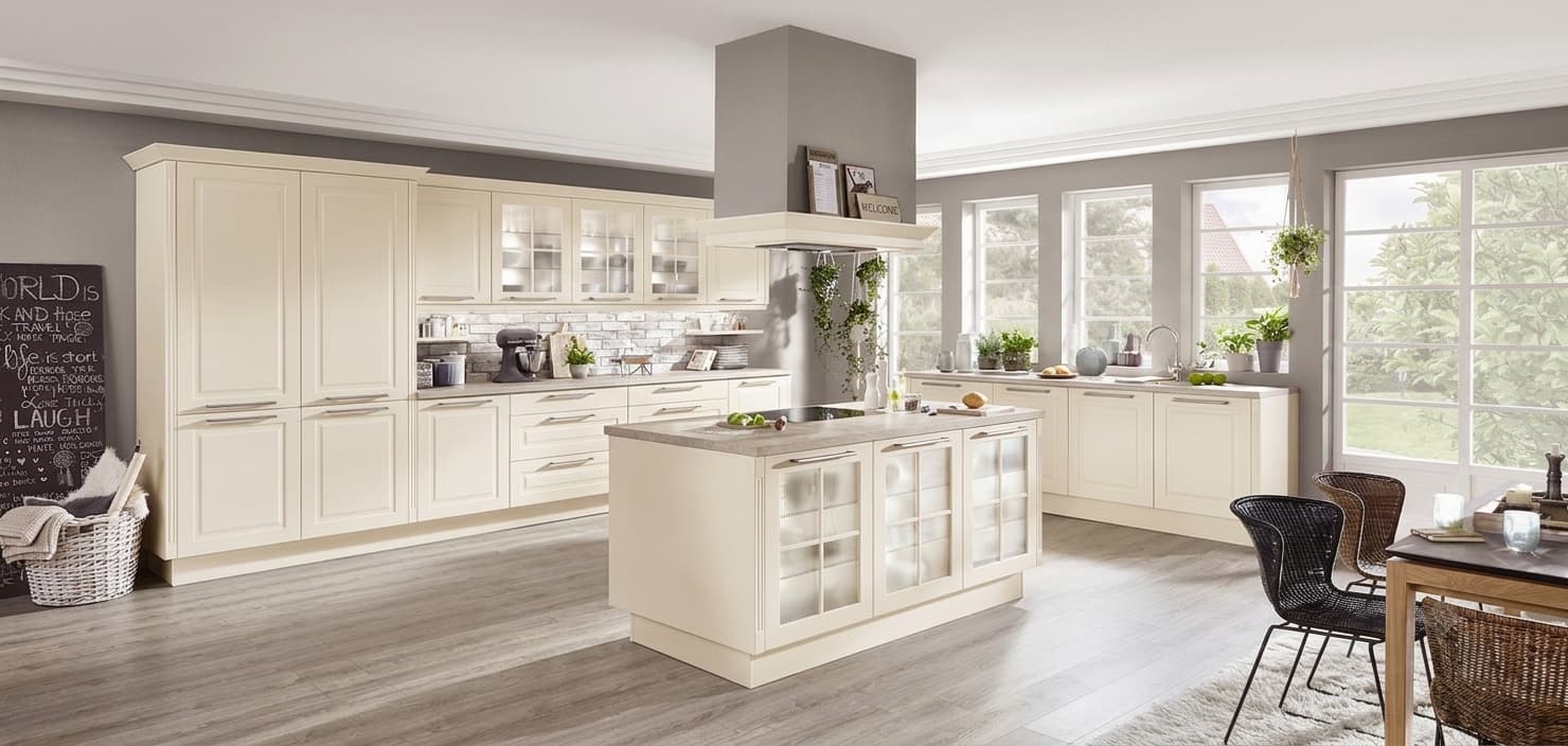 A spacious, modern cottage-style German kitchen featuring lacquer honed ivory shaker cabinets with classic glass-front display panes and slim bow handles, built around a central kitchen island with a matching limestone-reproduction worktop and an overhead chimney extractor hood.