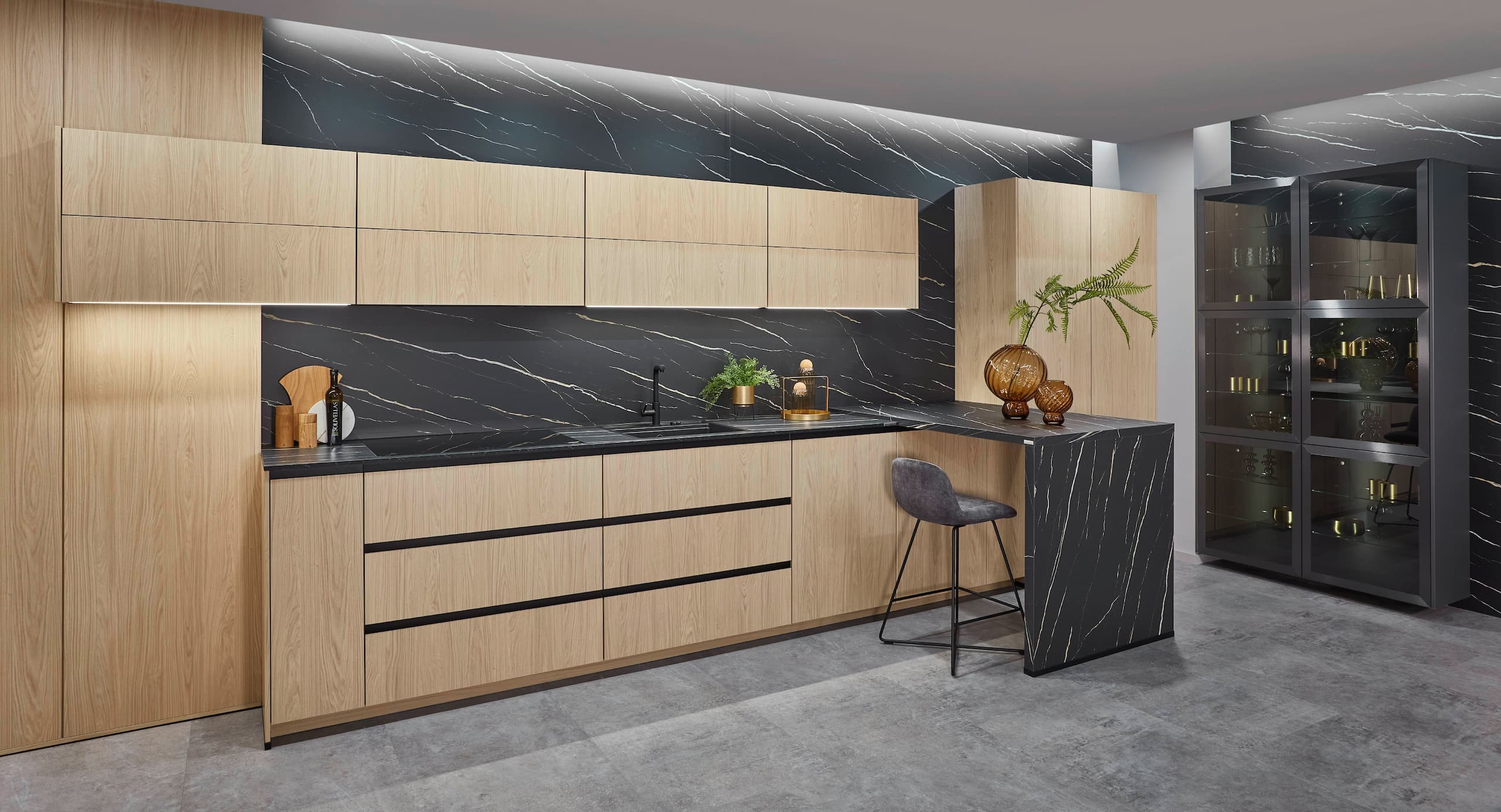 A spacious, modern German kitchen showcasing textured Bergamo oak reproduction base units and storage towers with minimalist vertical black handles, contrasted by matte sand-colored upper cabinetry, a matching drop-ceiling panel with integrated spotlights, and a large multi-compartment open wine rack island.
