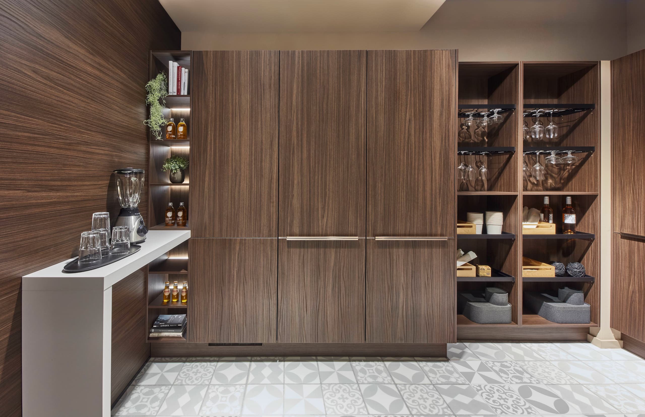 A sophisticated modern German kitchen displaying warm Milano walnut reproduction cabinetry with long, integrated horizontal metallic handles, matching wood-grain wall cladding, illuminated open storage niches, and two floor-to-ceiling open shelving units filled with glassware.