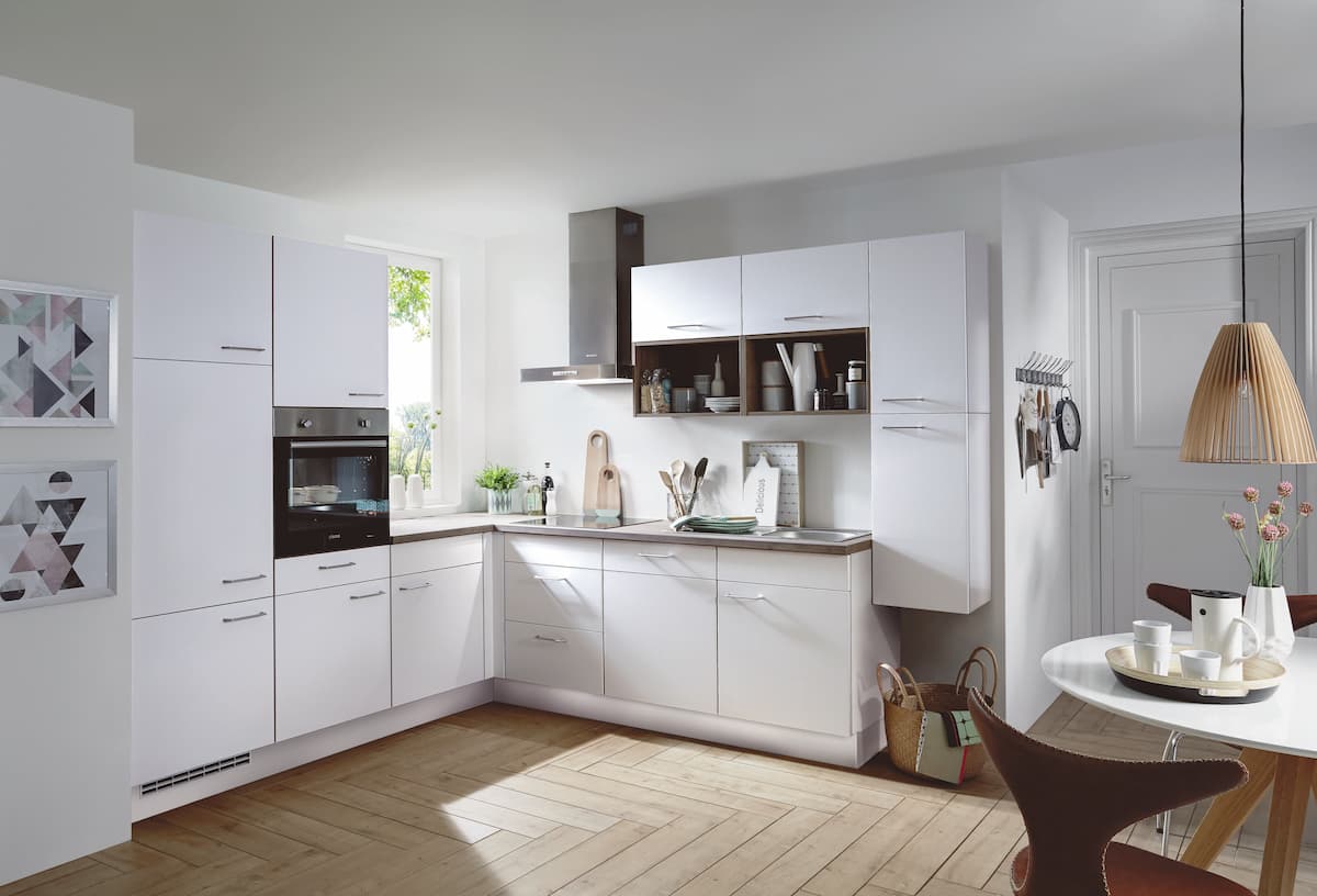 A modern L-shaped German kitchen featuring TOUCH 332 Alpine White supermatt cabinetry with sleek stainless steel bar handles, a warm wood-accented countertop and matching open shelving, an integrated stainless steel extractor hood, and a bright dining area with a wooden pendant light.